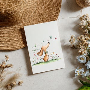 May include: A watercolour illustration of two dachshund dogs, one looking up at butterflies. The artwork is on a white card, surrounded by flowers, a straw hat, and a ball of twine. The dogs are brown with green accents.