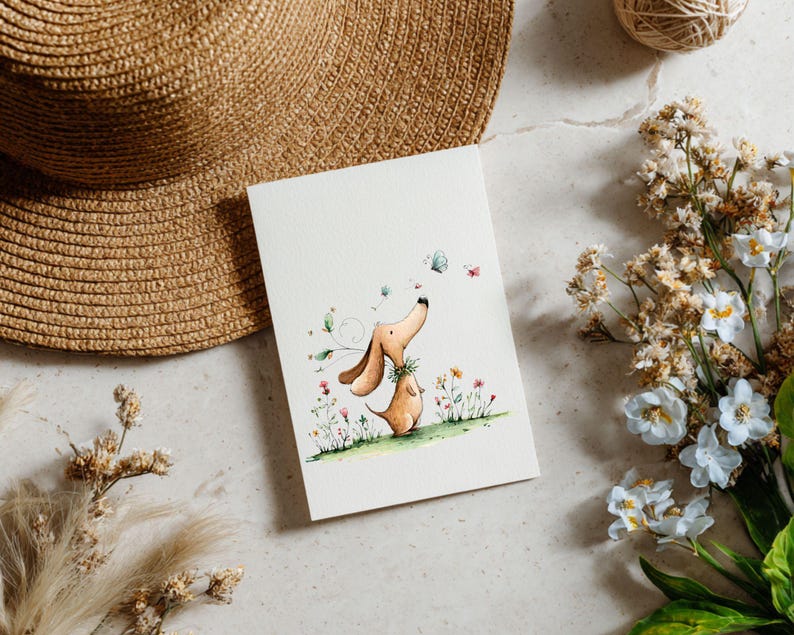 May include: A watercolour illustration of two dachshund dogs, one looking up at butterflies. The artwork is on a white card, surrounded by flowers, a straw hat, and a ball of twine. The dogs are brown with green accents.