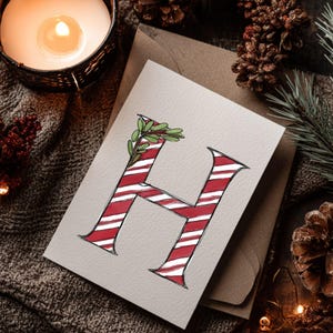 May include: A festive greeting card displaying a red and white striped letter 'H' adorned with green leaves. The card rests on a neutral surface, accompanied by pine cones, foliage, and a lit candle.