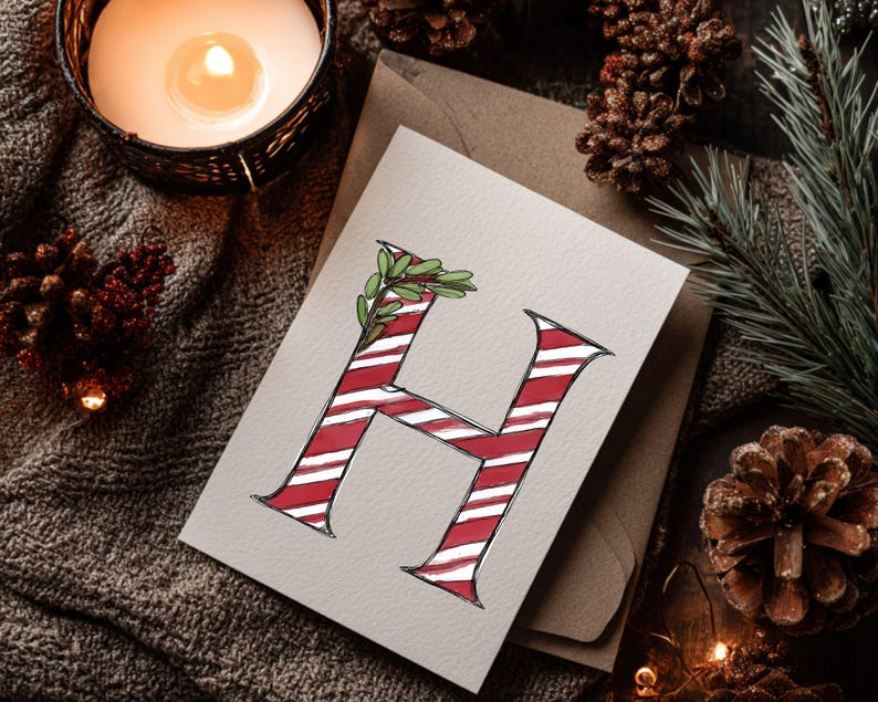 May include: A festive greeting card displaying a red and white striped letter 'H' adorned with green leaves. The card rests on a neutral surface, accompanied by pine cones, foliage, and a lit candle.