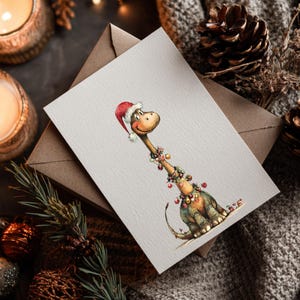 May include: A watercolor Christmas card featuring a friendly dinosaur wearing a Santa hat and festive garland. The card is on a neutral background with a brown envelope, pine cones, and candles, creating a cozy holiday scene.