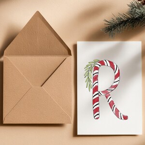 May include: A Christmas-themed greeting card with a candy cane-style letter 'R' in red and white stripes, decorated with green foliage. A brown envelope sits open beside the card. A sprig of evergreen is in the background.
