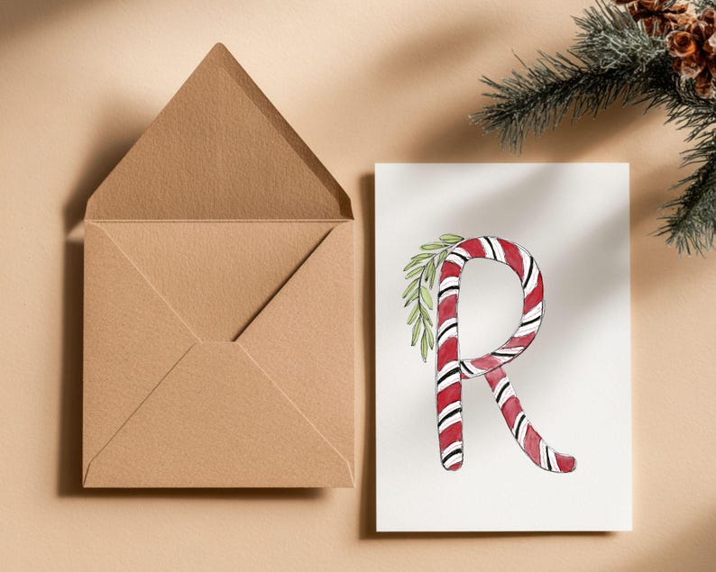 May include: A Christmas-themed greeting card with a candy cane-style letter 'R' in red and white stripes, decorated with green foliage. A brown envelope sits open beside the card. A sprig of evergreen is in the background.