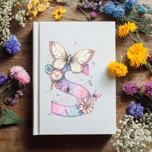 May include: A white journal with a watercolor illustration of a butterfly and the letter "S". The butterfly is cream-colored with pink accents, perched atop a pastel-colored letter "S" adorned with flowers and other butterflies. The journal is surrounded by colorful flowers.