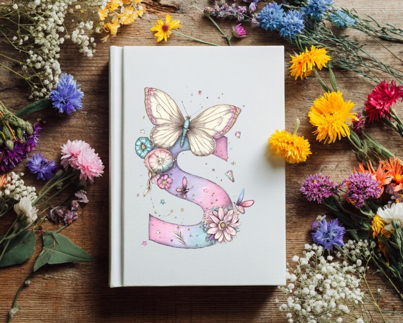 May include: A white journal with a watercolor illustration of a butterfly and the letter "S". The butterfly is cream-colored with pink accents, perched atop a pastel-colored letter "S" adorned with flowers and other butterflies. The journal is surrounded by colorful flowers.