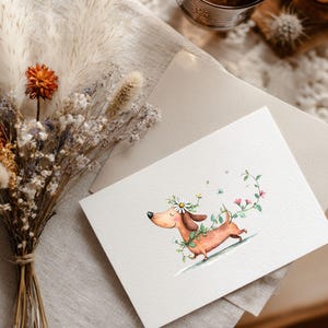 May include: A watercolour illustration of a dachshund dog adorned with a daisy crown and floral details. The dog is depicted running on a gentle slope, encircled by a green vine with pink flowers. The image is on a white card with a beige envelope, beside a bouquet of dried flowers.