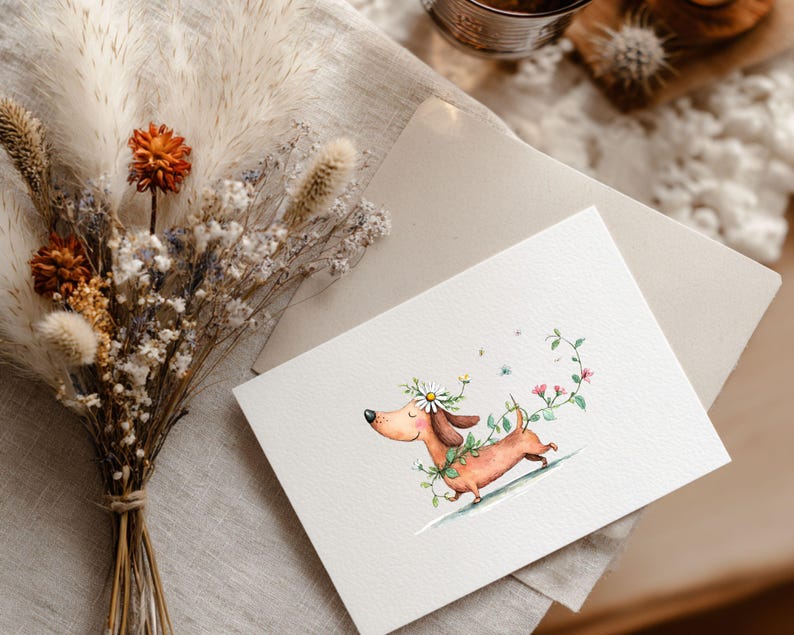 May include: A watercolour illustration of a dachshund dog adorned with a daisy crown and floral details. The dog is depicted running on a gentle slope, encircled by a green vine with pink flowers. The image is on a white card with a beige envelope, beside a bouquet of dried flowers.