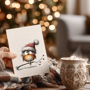 May include: A greeting card featuring a watercolor illustration of a cartoon robin wearing a Santa hat, perched on a branch with berries. A mug of hot beverage with whipped cream sits nearby. The background is blurred with Christmas lights.