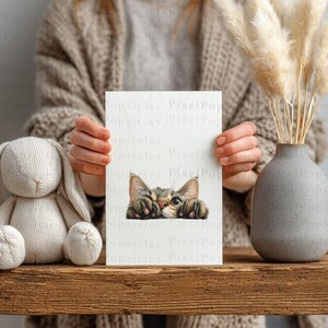 May include: A white card with a watercolour illustration of a tabby cat peeking over the edge. The card is held in front of a person. A stuffed bunny and a grey vase with dried pampas grass are on a wooden surface.