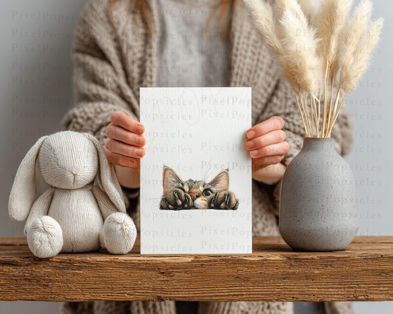 May include: A white card with a watercolour illustration of a tabby cat peeking over the edge. The card is held in front of a person. A stuffed bunny and a grey vase with dried pampas grass are on a wooden surface.