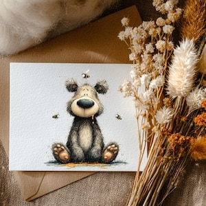 May include: A watercolour illustration of a bear with bees. The bear is sitting with honey dripping from its mouth. The image is on a white card with a brown envelope behind it. Dried flowers are in the background.