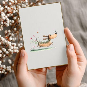 May include: A greeting card featuring a cartoon dachshund dog running. The dog is depicted in brown and tan with a green garland. The card has a gold border and is held by a person. The illustration is set against a white background.