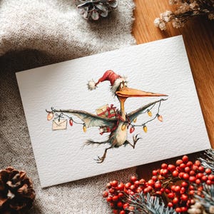 May include: A Christmas card featuring a cartoon pterodactyl wearing a Santa hat and carrying gifts and string lights. The pterodactyl has a long beak and is depicted in flight. The card is on a white background with holiday decorations.