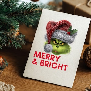 Peut inclure: Carte de No&euml;l avec un personnage vert portant un bonnet de P&egrave;re No&euml;l rouge et argent&eacute;. La carte indique "MERRY & BRIGHT" en texte rouge. La carte est sur une surface en bois avec des d&eacute;corations de No&euml;l.