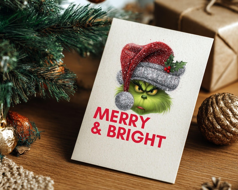 Peut inclure: Carte de No&euml;l avec un personnage vert portant un bonnet de P&egrave;re No&euml;l rouge et argent&eacute;. La carte indique "MERRY & BRIGHT" en texte rouge. La carte est sur une surface en bois avec des d&eacute;corations de No&euml;l.