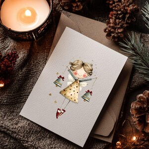 May include: A greeting card featuring a whimsical illustration of an angel holding bells. The angel wears a gold dress with heart patterns and red shoes. The card is set on a textured gray surface with a lit candle, pine cones, and greenery.
