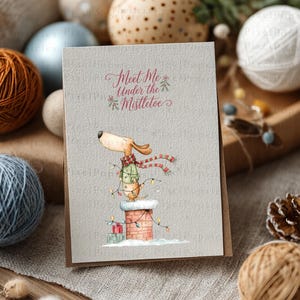 May include: A rectangular greeting card with a textured light gray surface featuring a watercolor illustration of a dog wearing a green sweater and red scarf, standing on a brick chimney with string lights and the text Meet Me Under the Mistletoe.