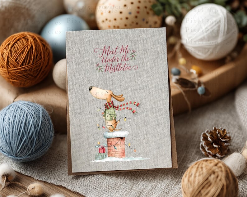 May include: A rectangular greeting card with a textured light gray surface featuring a watercolor illustration of a dog wearing a green sweater and red scarf, standing on a brick chimney with string lights and the text Meet Me Under the Mistletoe.