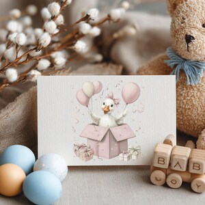 May include: A greeting card with a watercolour illustration of a duckling emerging from a pink gift box, holding pink balloons. The card is surrounded by a teddy bear, wooden toy train, and pastel-coloured eggs, creating a soft, baby-themed scene.
