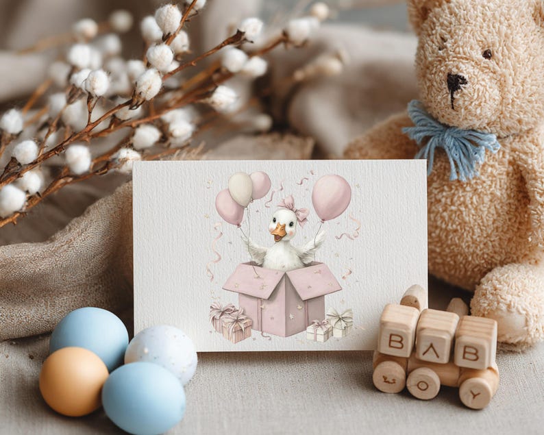 May include: A greeting card with a watercolour illustration of a duckling emerging from a pink gift box, holding pink balloons. The card is surrounded by a teddy bear, wooden toy train, and pastel-coloured eggs, creating a soft, baby-themed scene.