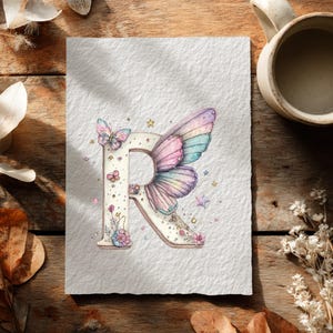 May include: Watercolor illustration of the letter "R" adorned with a butterfly wing design in pastel colors. The letter is decorated with butterflies, stars, and floral accents. The artwork is on textured paper, with a mug and dried flowers in the background.