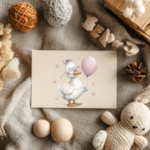 May include: A greeting card with a whimsical illustration of a white duck wearing glasses and a bow, holding a pink balloon. The card is surrounded by decorative items such as yarn balls, a wooden box, a pine cone, and a knitted bunny.