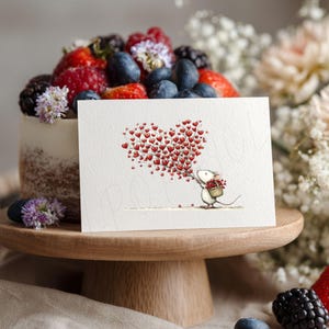 May include: A greeting card with a heart-shaped design made of red hearts, held by a cartoon mouse. The card sits on a wooden cake stand, with a cake topped with berries and flowers in the background.