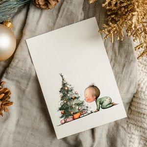Puede incluir: Una tarjeta navide&ntilde;a de acuarela con un beb&eacute; con un mono verde que se arrastra hacia un &aacute;rbol de Navidad decorado con regalos. La tarjeta est&aacute; sobre una superficie de lino neutro con adornos dorados y pi&ntilde;as.