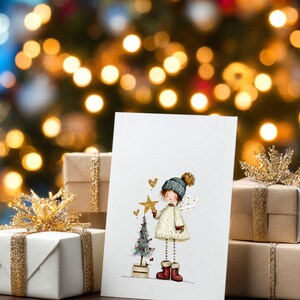May include: A Christmas card featuring an illustration of a girl angel holding a star, with a small Christmas tree. The card is propped up in front of wrapped gifts with gold ribbon and a blurred Christmas tree background.