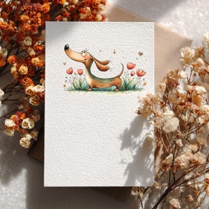 May include: A blank greeting card with a watercolour illustration of a dachshund dog. The dog has brown fur and a green belly, and is in a field of grass and flowers. The card is surrounded by dried flowers.