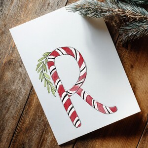 May include: A white card featuring a watercolour illustration of the letter "R" formed by a red and white striped candy cane. Green leaves are attached. The card rests on a wooden surface, with a pine branch in the background.