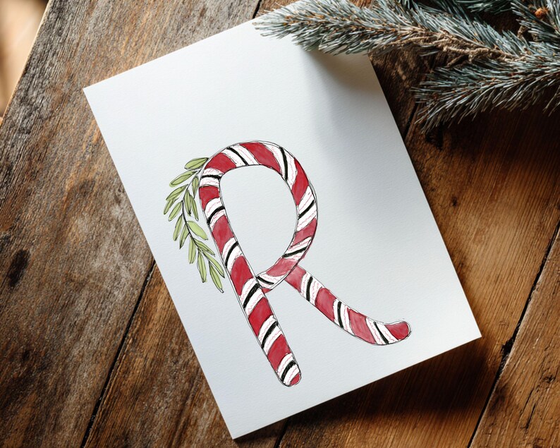 May include: A white card featuring a watercolour illustration of the letter "R" formed by a red and white striped candy cane. Green leaves are attached. The card rests on a wooden surface, with a pine branch in the background.