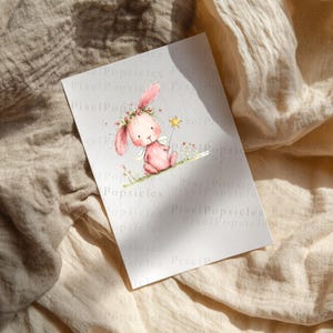 Op de afbeelding: Een rechthoekige witte kaart met een aquarelillustratie van een roze konijn met een bloemenkrans en kleine witte vleugels. Het konijn houdt een gele sterrenstaf vast en zit op een dunne groene lijn. De tekst PixelPopsicles is zichtbaar.
