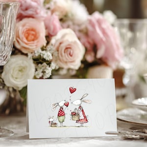 May include: A greeting card featuring a whimsical illustration of two rabbits holding hearts, with a smaller rabbit family nearby. The rabbits are dressed in red and green outfits, with a red heart floating above them. The card is set on a table with flowers and glassware.