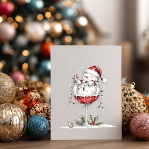 Peut inclure: Une carte de v&oelig;ux rectangulaire pr&eacute;sentant une illustration &agrave; l'encre et &agrave; l'aquarelle d'un bonhomme de neige souriant portant un bonnet de P&egrave;re No&euml;l rouge, assis dans un grand verre &agrave; vin rempli de liquide rouge, orn&eacute; de guirlandes lumineuses.