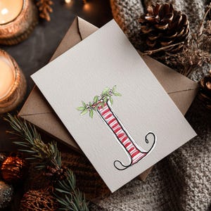 May include: A greeting card with a watercolour illustration of the letter "I", striped in red and white, decorated with green leaves and berries. The card rests on a neutral surface with a brown envelope, pine cones, and candles.