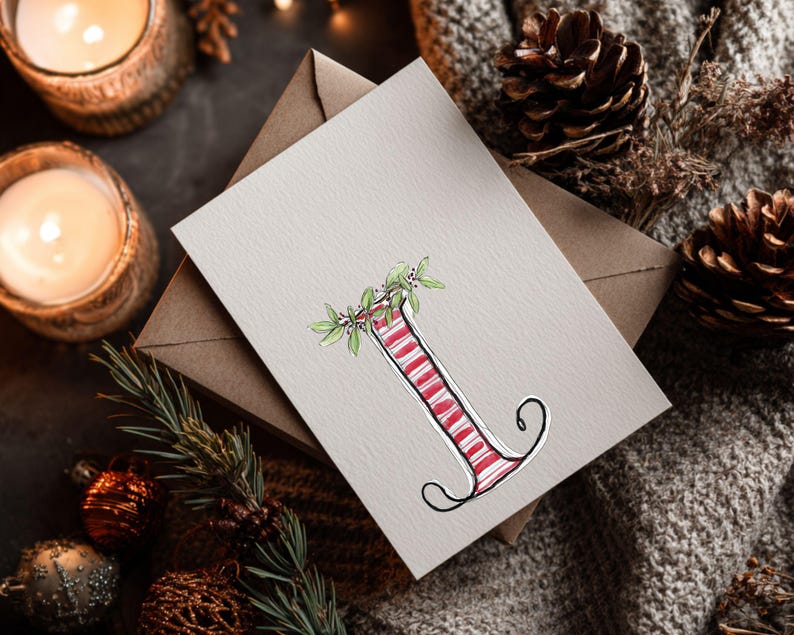 May include: A greeting card with a watercolour illustration of the letter "I", striped in red and white, decorated with green leaves and berries. The card rests on a neutral surface with a brown envelope, pine cones, and candles.