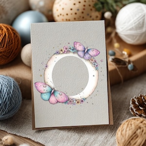 May include: A greeting card with a light gray background features a circular frame with a butterfly design. The butterflies have pink and blue wings. The frame is decorated with small flowers. The card is on a brown envelope.