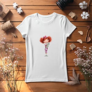 Puede incluir: Camiseta blanca con una ilustraci&oacute;n de dibujos animados de una enfermera con cabello rizado rojo, vestida con un uniforme rosa y sosteniendo un portapapeles. El dise&ntilde;o est&aacute; centrado en la camiseta, ofreciendo una est&eacute;tica caprichosa y de tem&aacute;tica m&eacute;dica.
