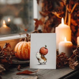 May include: A greeting card featuring a watercolor illustration of a small, fluffy animal carrying a large red apple. The card is propped up in front of a pumpkin and lit candles, creating a cozy autumn scene.