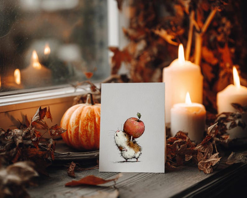 May include: A greeting card featuring a watercolor illustration of a small, fluffy animal carrying a large red apple. The card is propped up in front of a pumpkin and lit candles, creating a cozy autumn scene.