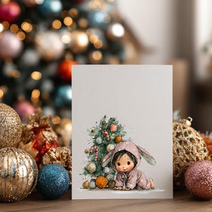 Puede incluir: Tarjeta de felicitaci&oacute;n navide&ntilde;a con una ilustraci&oacute;n en acuarela de un beb&eacute; con un traje de conejito rosa, cerca de un &aacute;rbol de Navidad decorado. La tarjeta est&aacute; colocada sobre un fondo de adornos navide&ntilde;os borrosos y una superficie de madera.