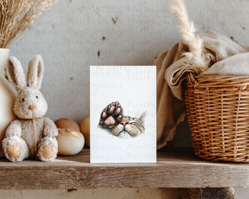 May include: A watercolour print featuring a sleeping cat, its paw raised. The cat's face has brown and grey stripes, and the paw has pink pads. The print is on a wooden shelf with a stuffed bunny and a wicker basket.