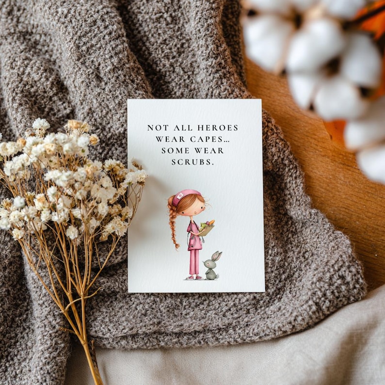 Puede incluir: Una tarjeta de felicitaci&oacute;n con el texto "NOT ALL HEROES WEAR CAPES... SOME WEAR SCRUBS." presenta una ilustraci&oacute;n de una enfermera y un conejo. La tarjeta est&aacute; sobre una manta gris con flores secas.