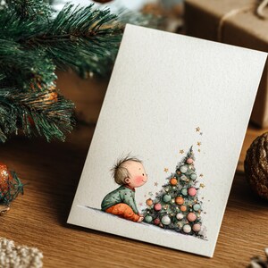Puede incluir: Una tarjeta de Navidad con una ilustraci&oacute;n en acuarela de un beb&eacute; con ropa verde y naranja, mirando un &aacute;rbol de Navidad decorado. La tarjeta est&aacute; sobre una superficie de madera con adornos navide&ntilde;os.
