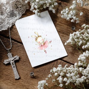Op de afbeelding: Een rechthoekige witte kaart met een aquarelillustratie van een konijn als ballerina in een roze tutu met een bloemenkrans. Naast de kaart ligt een zilverkleurige crucifix-hanger aan een bijpassende ketting, omringd door witte gipskruidbloemen.