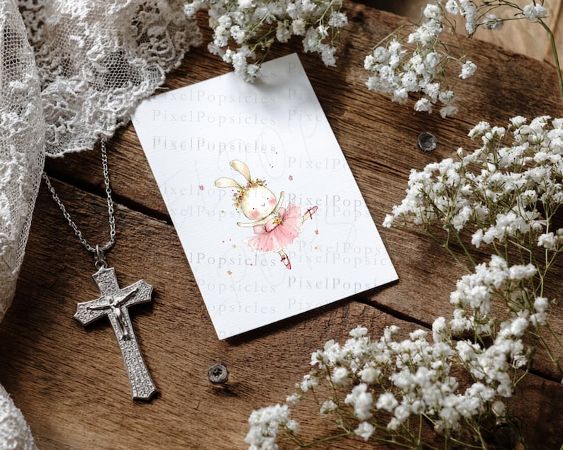 Op de afbeelding: Een rechthoekige witte kaart met een aquarelillustratie van een konijn als ballerina in een roze tutu met een bloemenkrans. Naast de kaart ligt een zilverkleurige crucifix-hanger aan een bijpassende ketting, omringd door witte gipskruidbloemen.