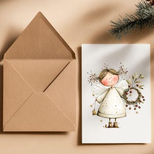 May include: A greeting card featuring a whimsical angel illustration, holding a wreath and a lantern. The angel is dressed in a cream-colored dress with gold stars. A brown envelope is next to the card. A sprig of evergreen is in the background.