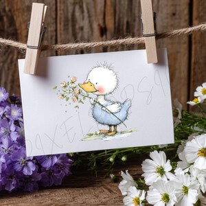 May include: A greeting card featuring a cartoon duckling in a blue outfit, holding flowers. The card is clipped to a clothesline with a wooden clothespin. The background includes purple and white flowers and a wooden surface.