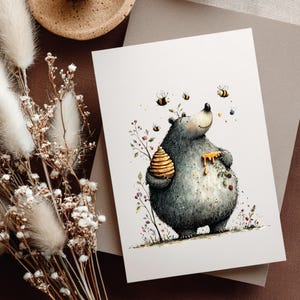 May include: A watercolour illustration of a bear with a beehive and honey dripping down its chest. Bees fly around the bear, and the artwork is on a white card. The card is on a brown surface with dried flowers.